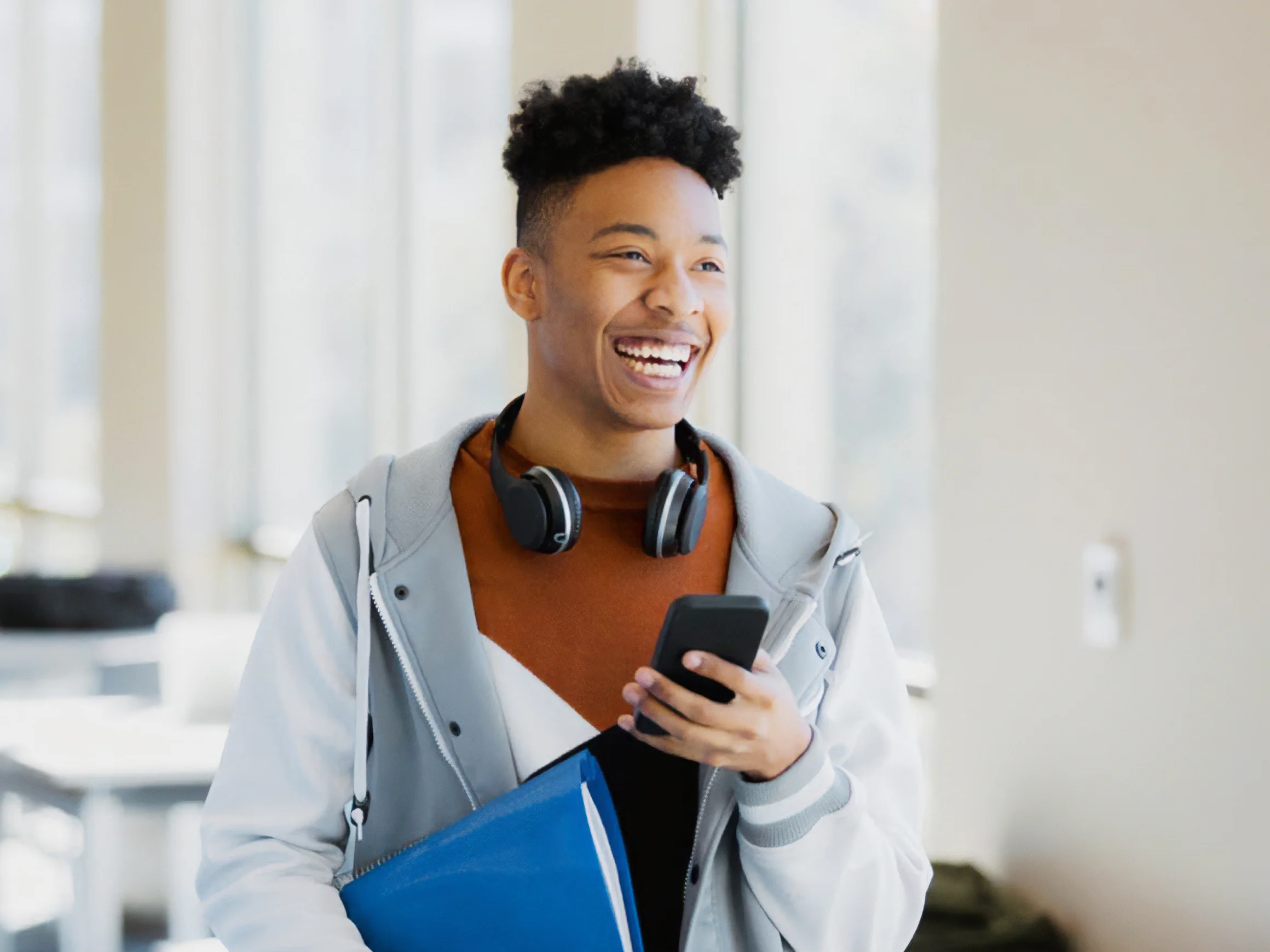 NDIS Provider Wollongong-A happy man holding a smartphone in one hand and a blue folder in the other. The person is wearing a light gray jacket over a brown shirt and has a pair of headphones around their neck.