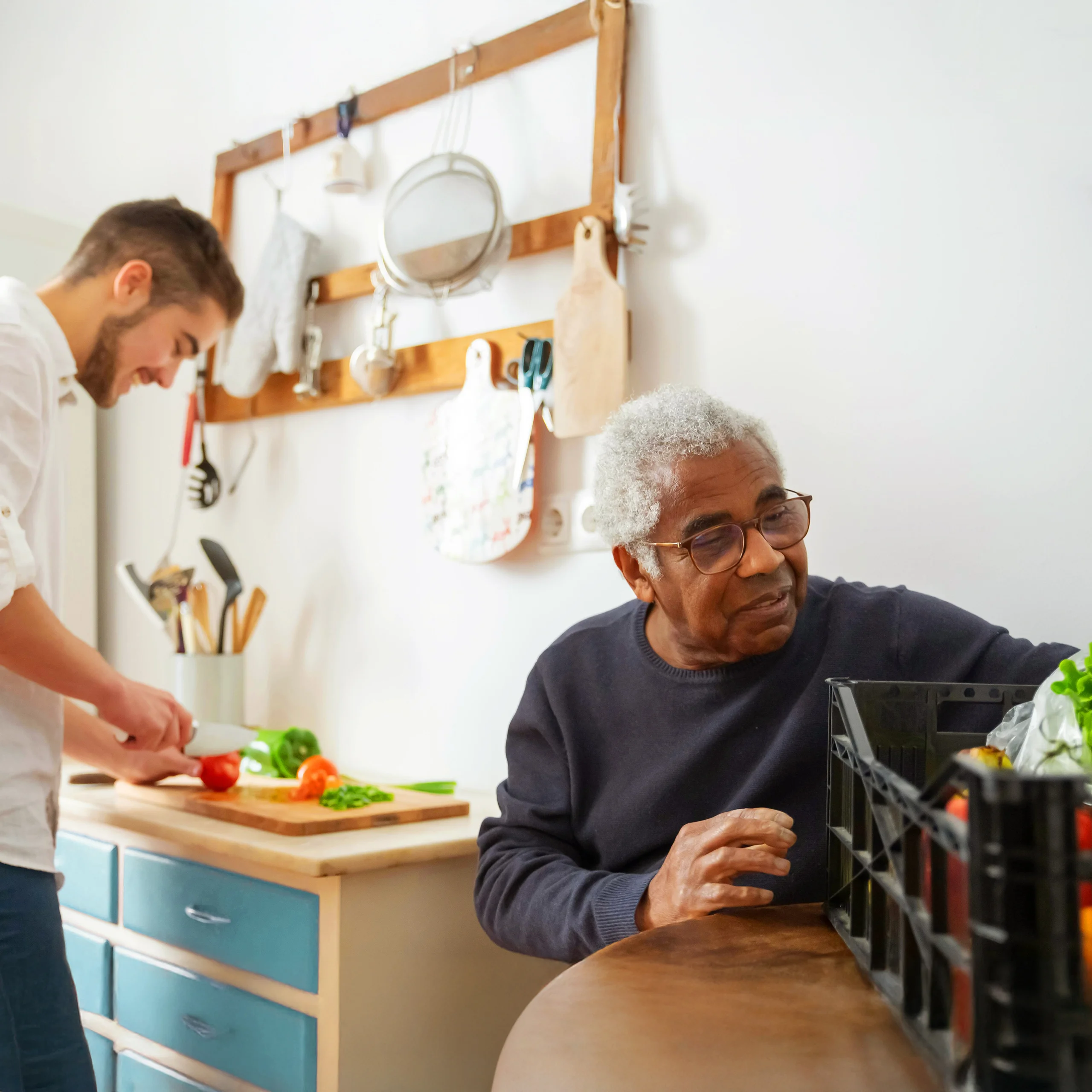 A man standing at a kitchen counter, chopping vegetables, including a red bell pepper and green leafy vegetables and is helping the old man with meal preparation or cooking