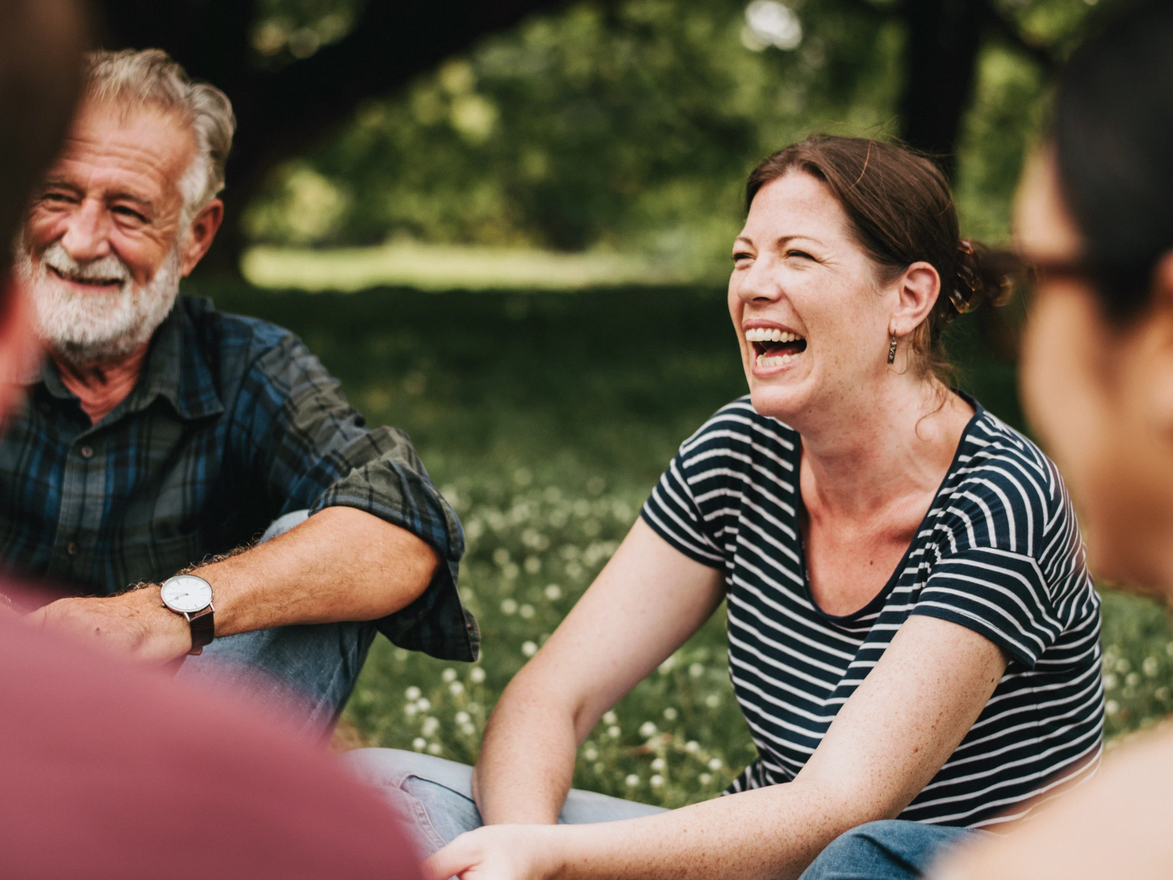 The image shows a group of people sitting on a grassy area outdoors, likely in a park, surrounded by trees and greenery. Everyone is sitting in a circle, chatting and smiling, radiating happiness and enjoying each other's company in a relaxed setting.